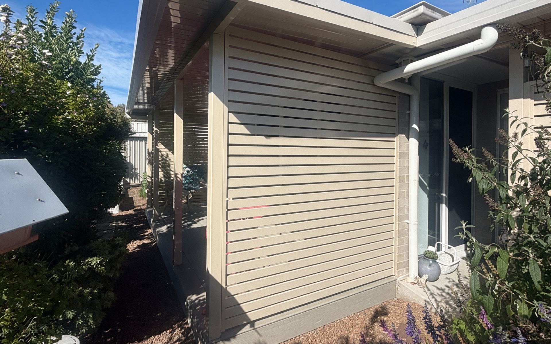 Tan house exterior with a window air conditioner, side fence, and garden path in bright sunlight