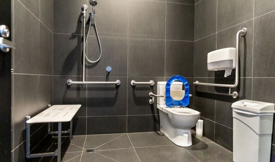 Accessible bathroom with black tile, toilet with blue seat cover, grab bars, shower, sink, and trash can.