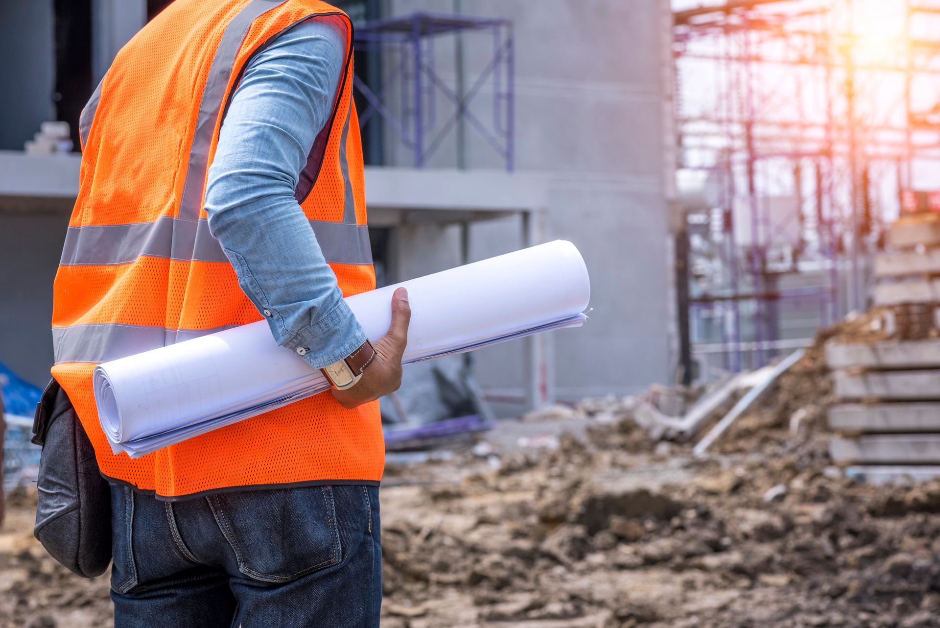 Operaio edile con giubbotto di sicurezza arancione che tiene in mano dei progetti arrotolati in un cantiere.
