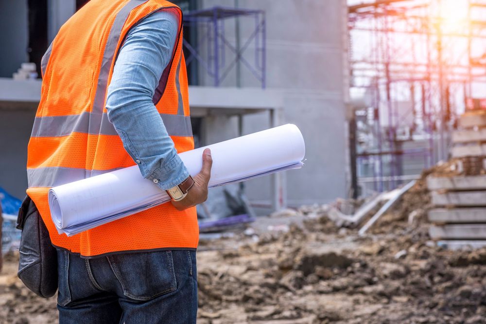 Operaio edile con giubbotto di sicurezza arancione che tiene in mano dei progetti arrotolati in un cantiere.