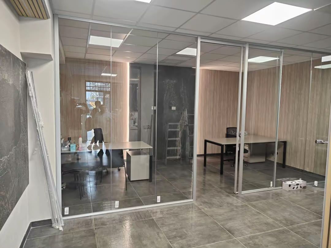 Ufficio vuoto con pareti divisorie in vetro, pavimento in piastrelle grigie e un tavolo in fondo.