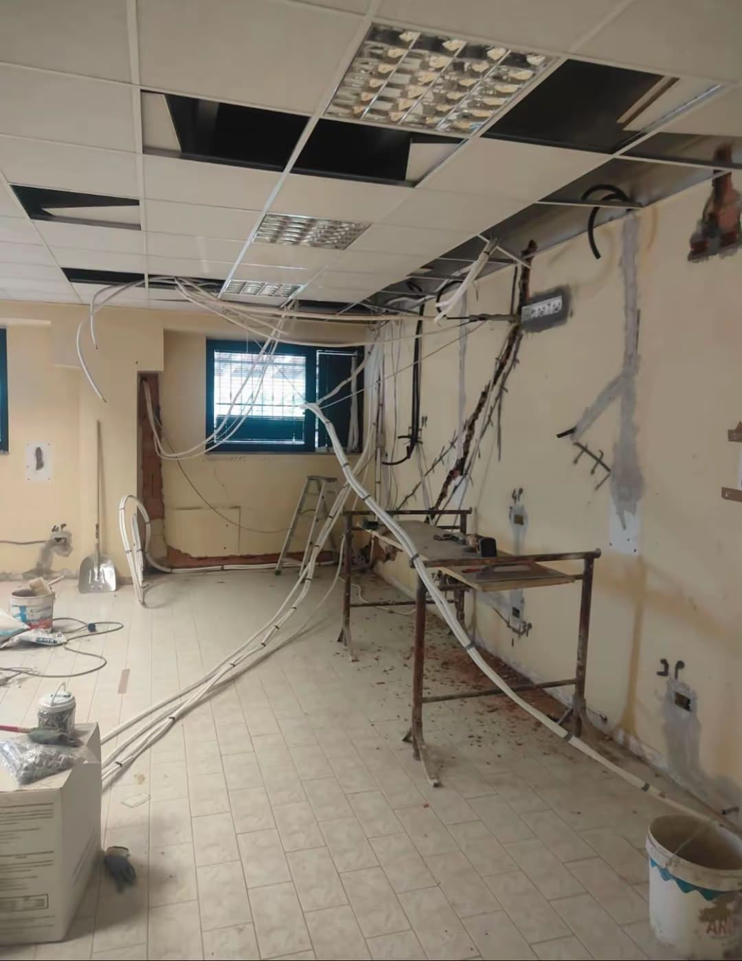 Interni di ufficio danneggiati con pannelli del soffitto rotti, fili elettrici scoperti e detriti sul pavimento.