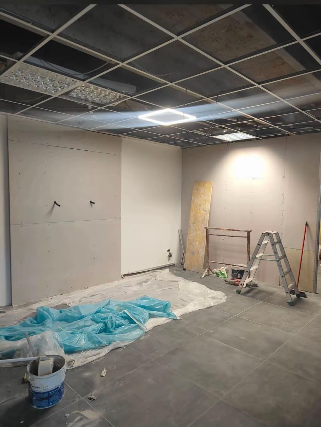 Stanza parzialmente ristrutturata con controsoffitto, scala, attrezzi e teli di plastica sul pavimento.
