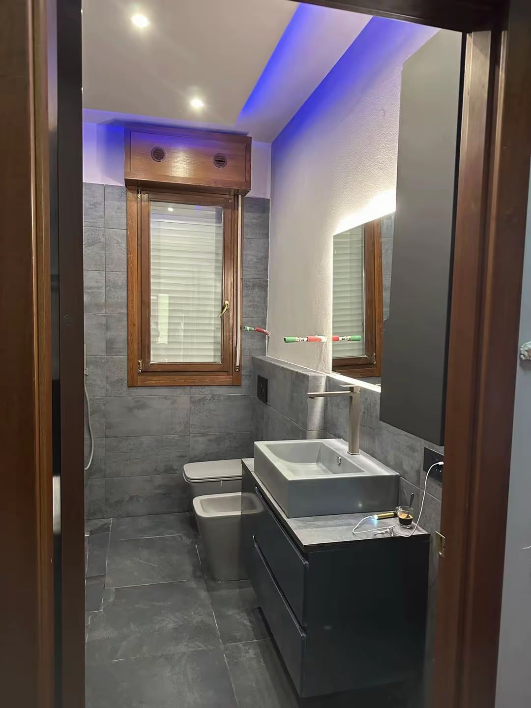 Bagno moderno con piastrelle scure, mobiletto nero, lavabo quadrato, specchio e illuminazione a soffitto blu.