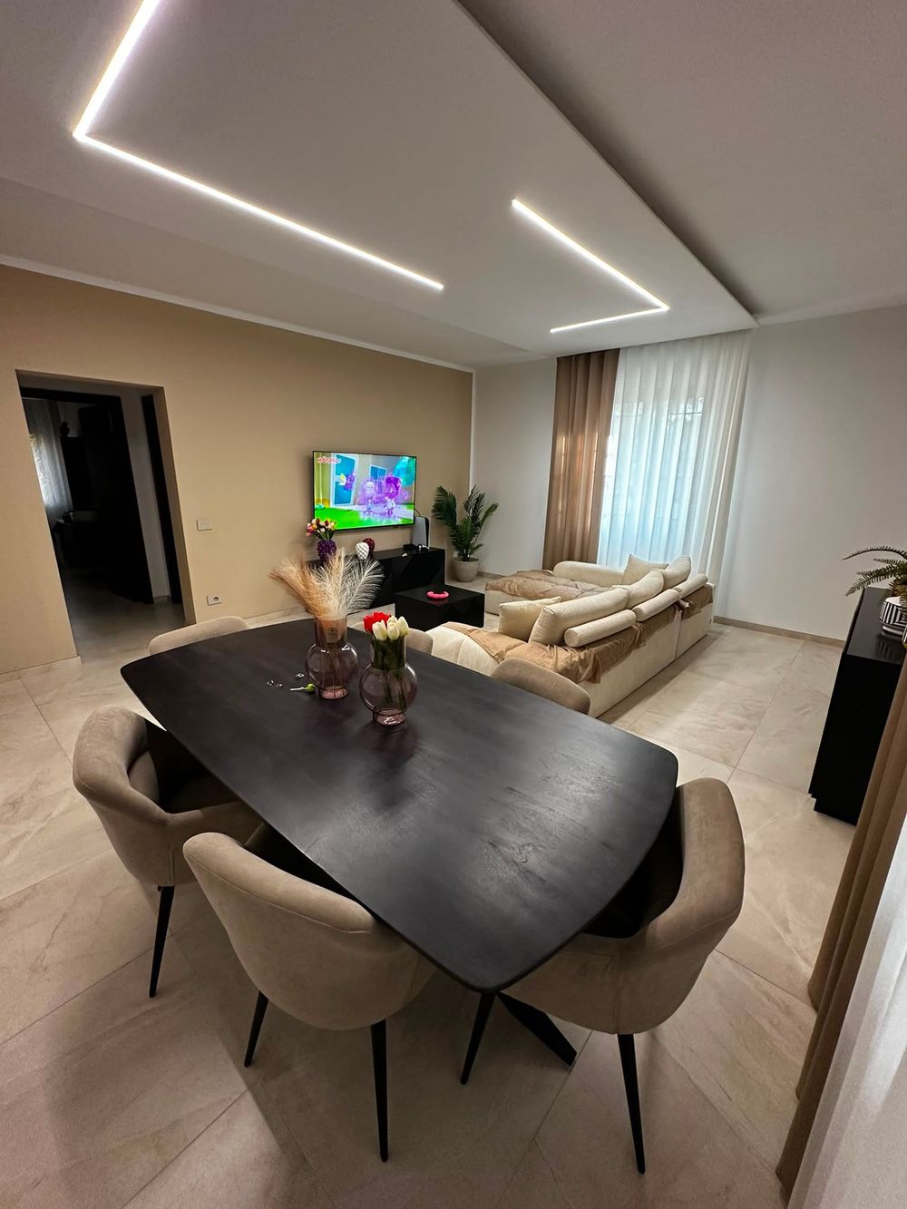 Soggiorno e sala da pranzo moderni con divani beige, tavolo da pranzo scuro, TV e faretti a incasso a soffitto.
