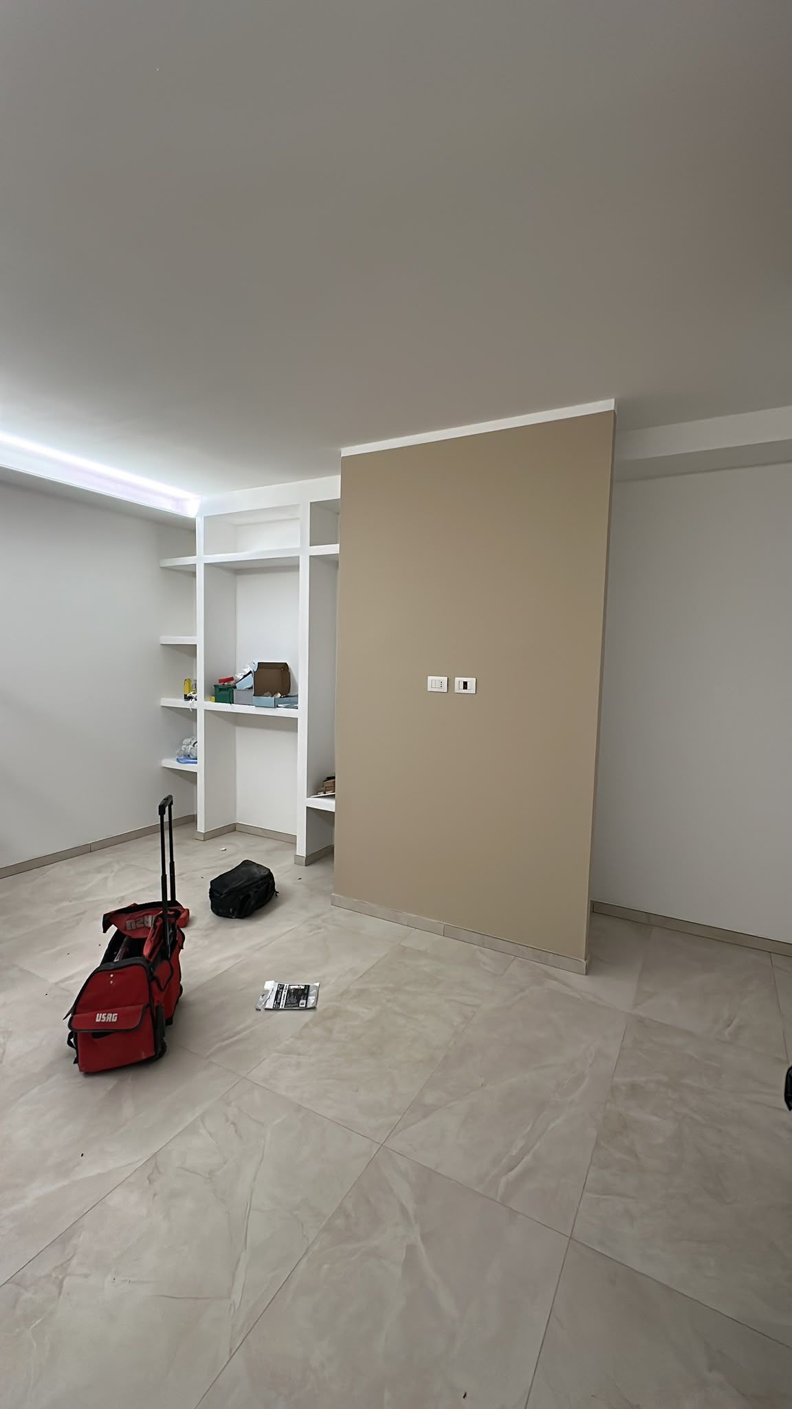 Stanza vuota con parete beige, scaffali a muro e una borsa rossa sul pavimento piastrellato.