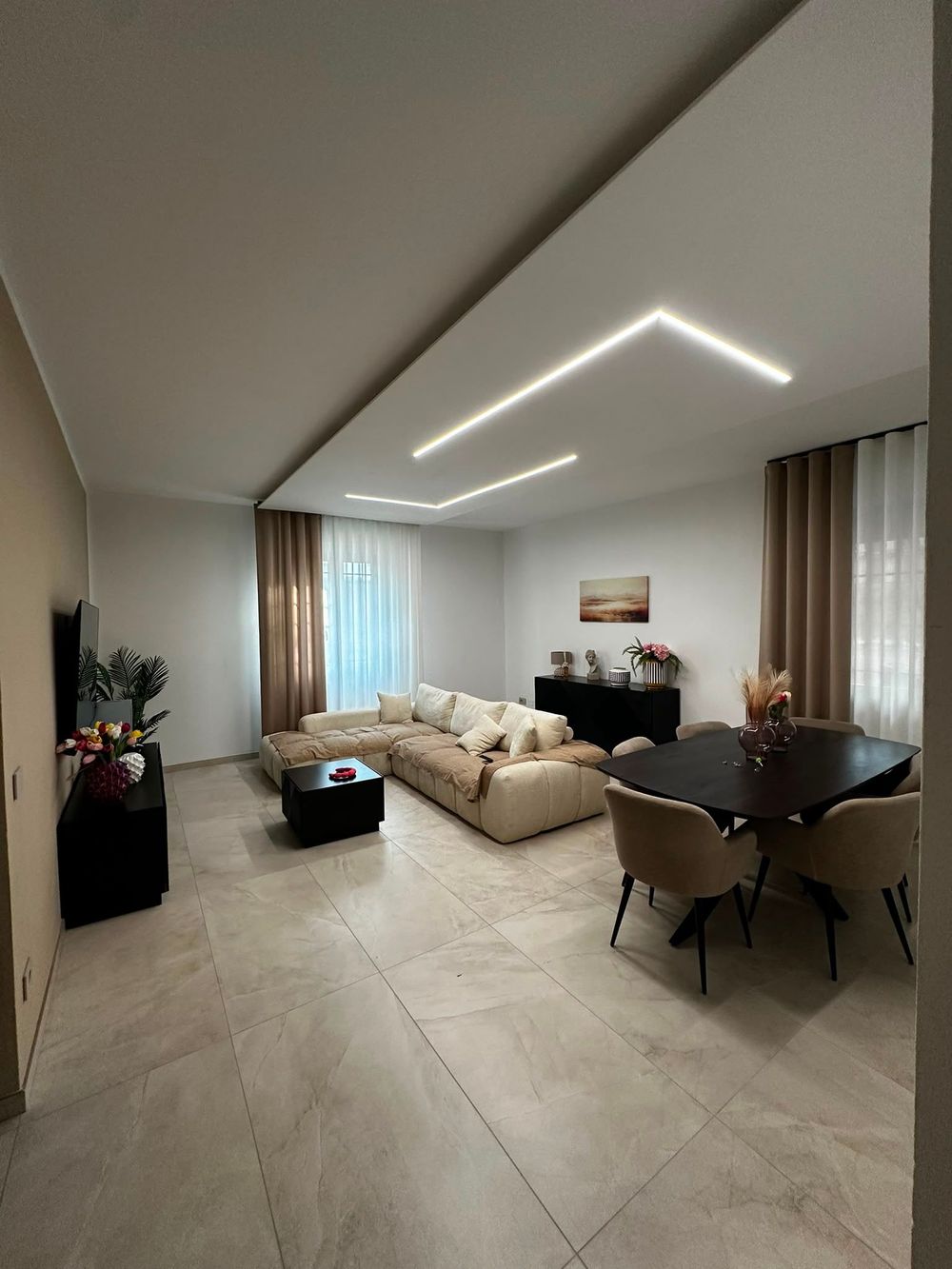 Soggiorno moderno con divano componibile beige, tavolo da pranzo, ampie finestre e lampade a soffitto geometriche.