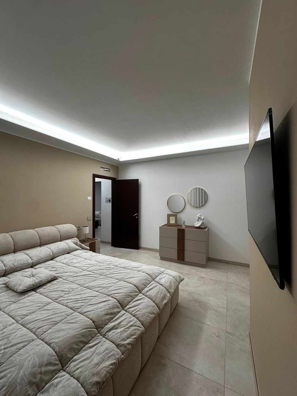 Camera da letto moderna con un grande letto, moquette beige, toletta e TV a parete in tonalità neutre.