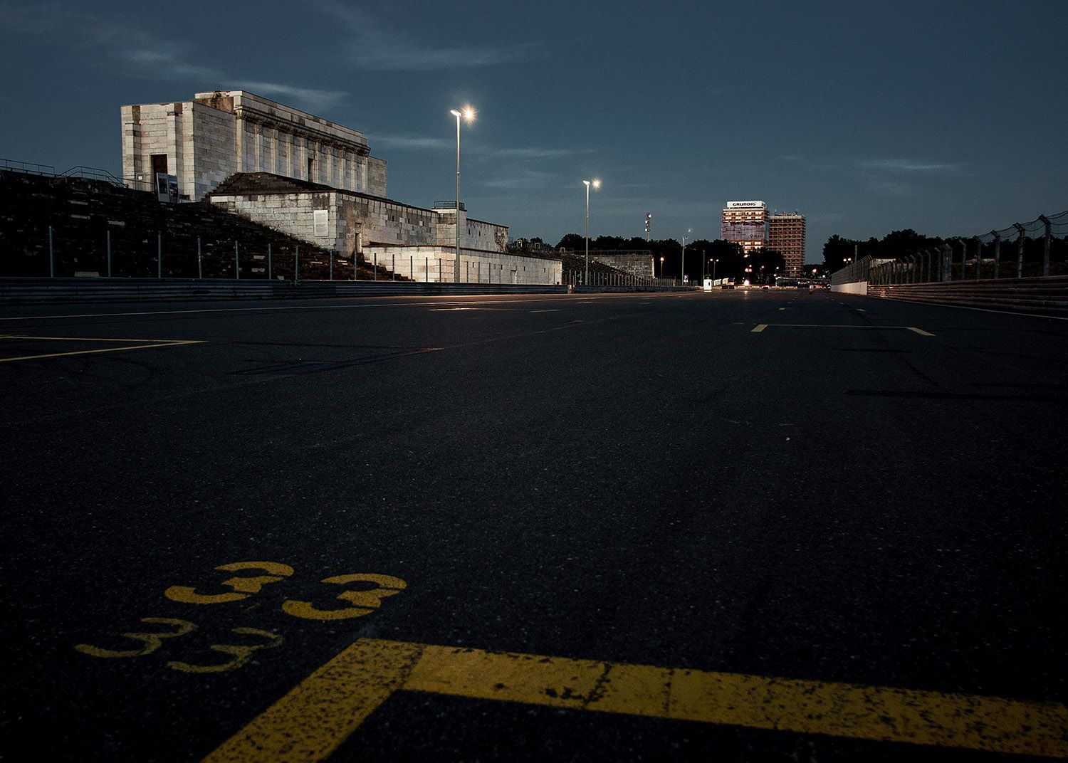Nachtelijk uitzicht op een parkeerplaats en een groot betonnen gebouw met straatverlichting onder een schemerige hemel.