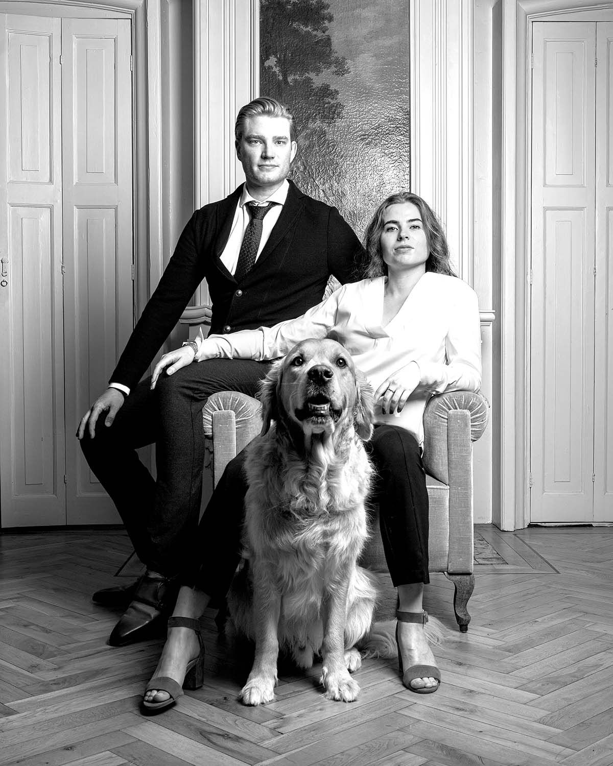 Een man in pak en een vrouw zitten met een golden retriever in een salon met luiken.