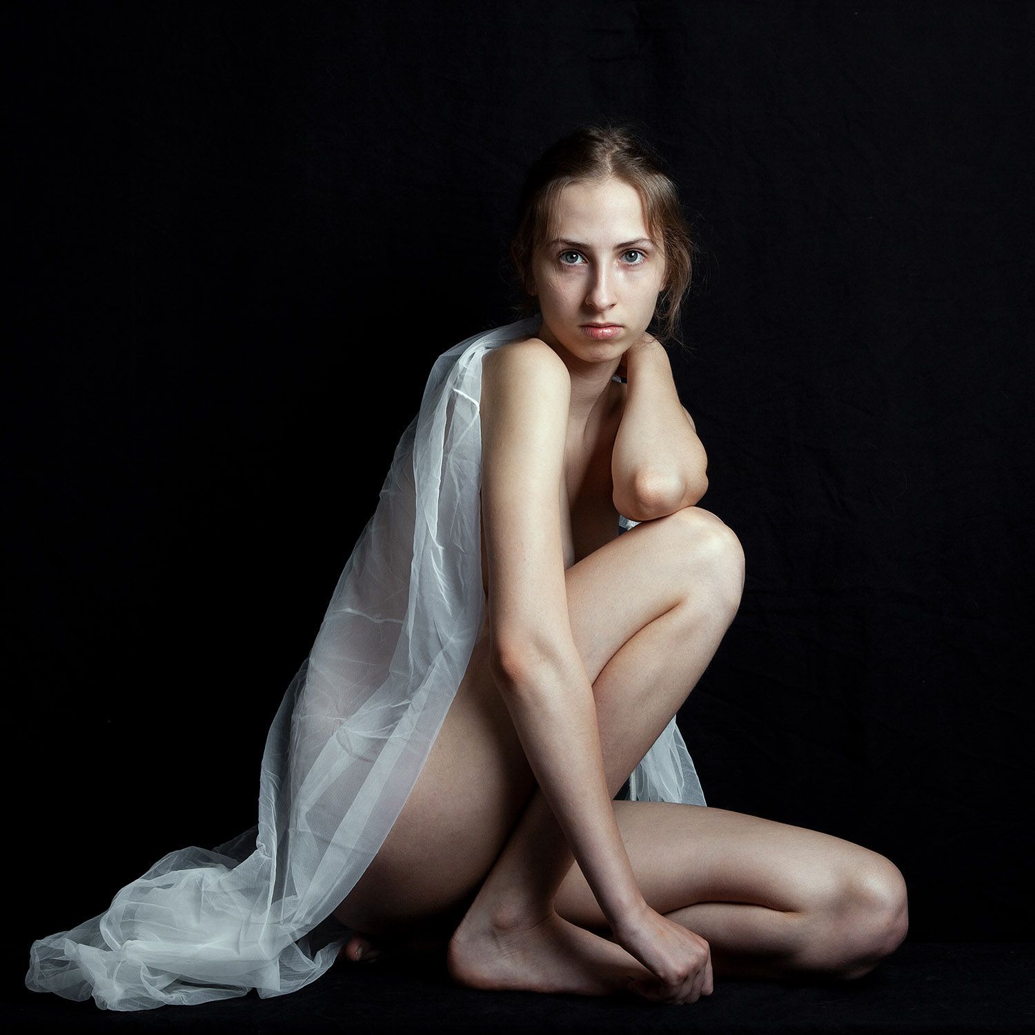 Vrouw in een doorschijnende witte stof, gehurkt tegen een zwarte achtergrond.