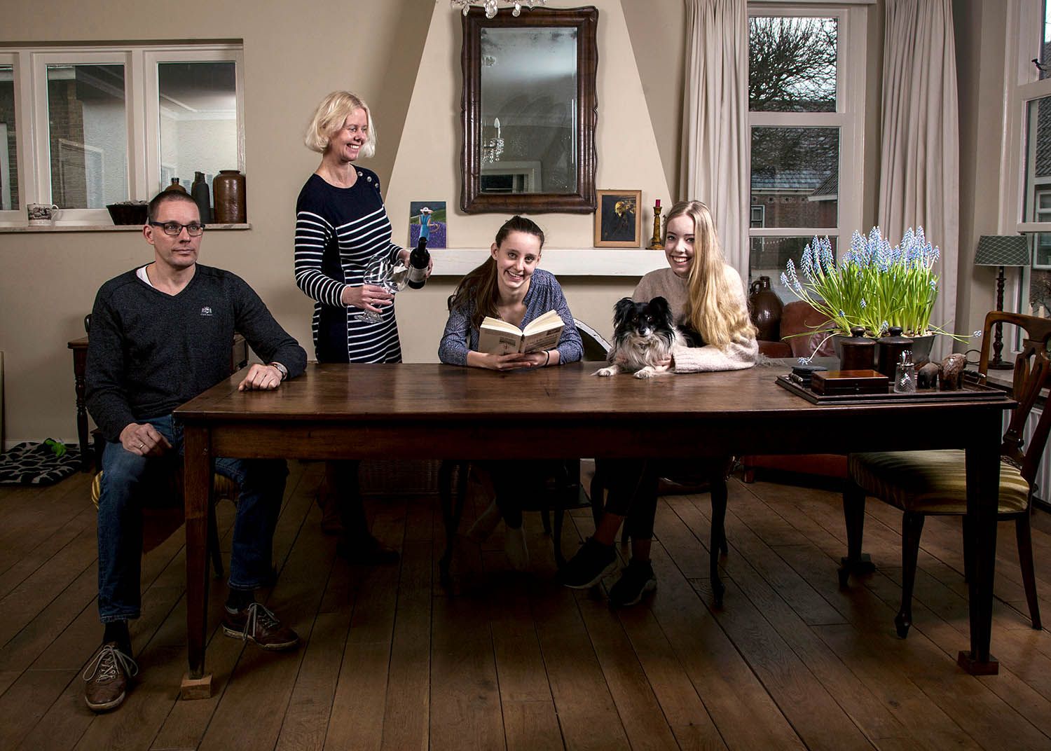Een gezin zit aan een houten tafel in de woonkamer, met een hond. Eén persoon leest, de anderen kijken toe.