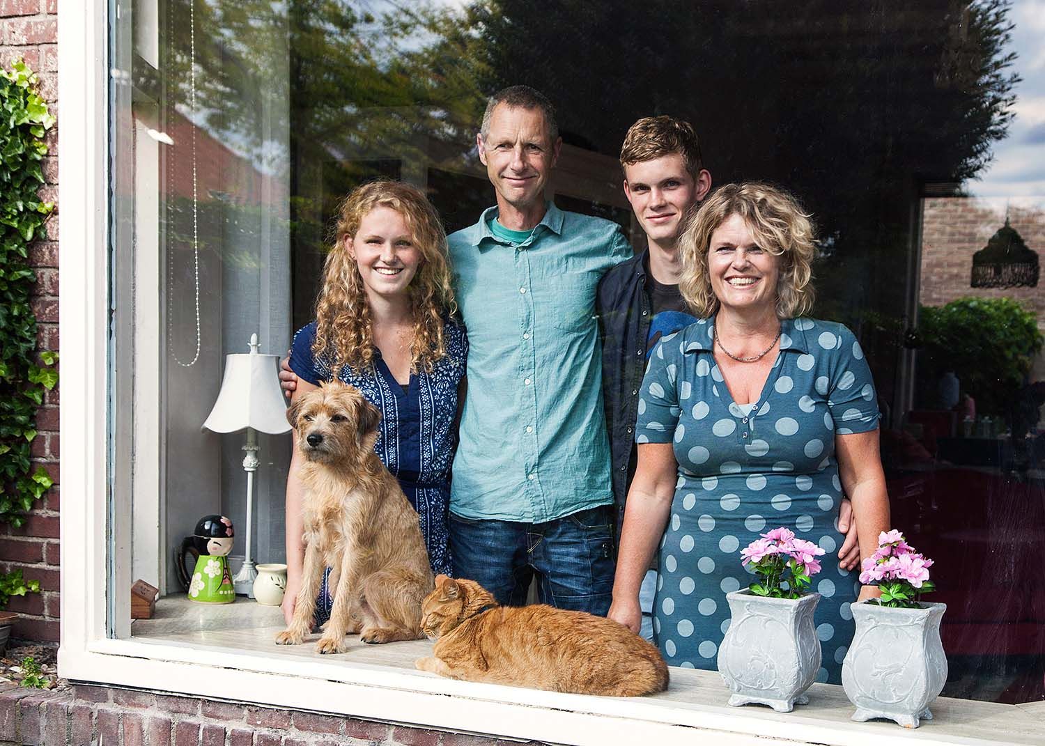 Een gezin van vier met twee honden poseert voor een raam; het is een zonnige dag.