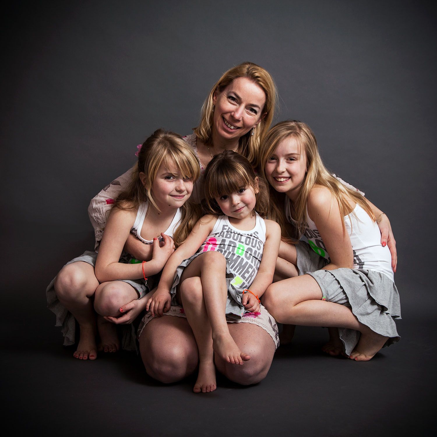 Een vrouw en drie meisjes, lachend, hurkend dicht bij elkaar, tegen een grijze achtergrond.