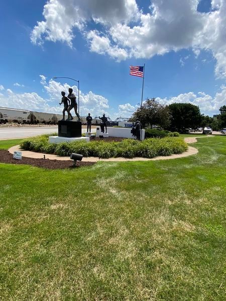 A statue of soldiers is in the middle of a lush green field with an american flag flying in the background.