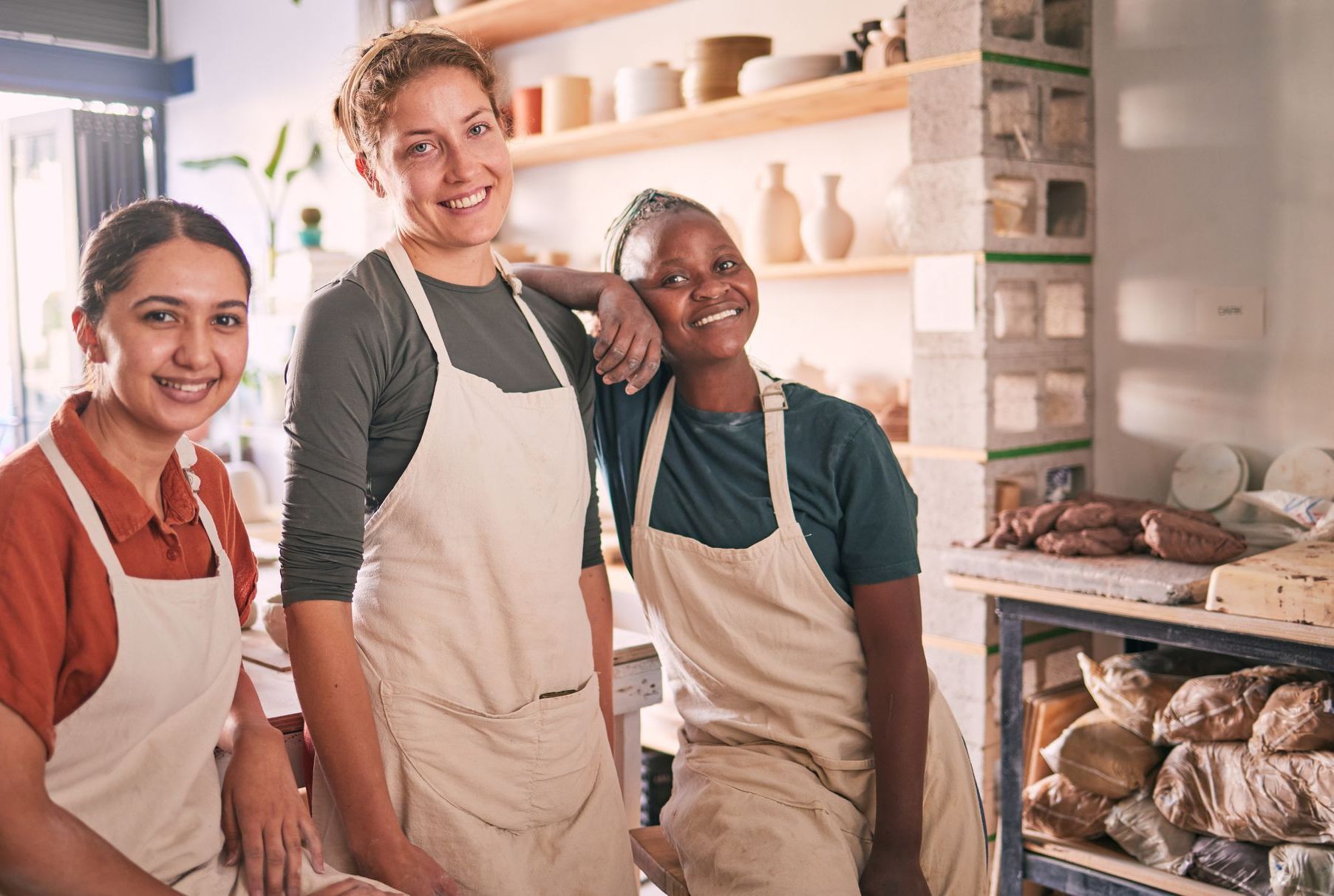 Three women in aprons are posing for a picture in a pottery studio.
