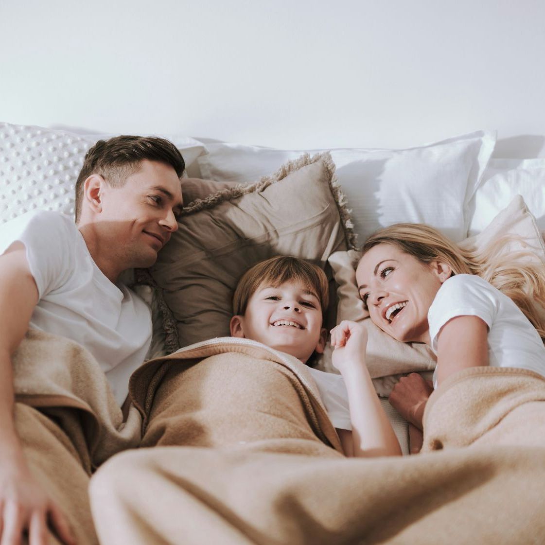 A family is laying in bed under a blanket and smiling.