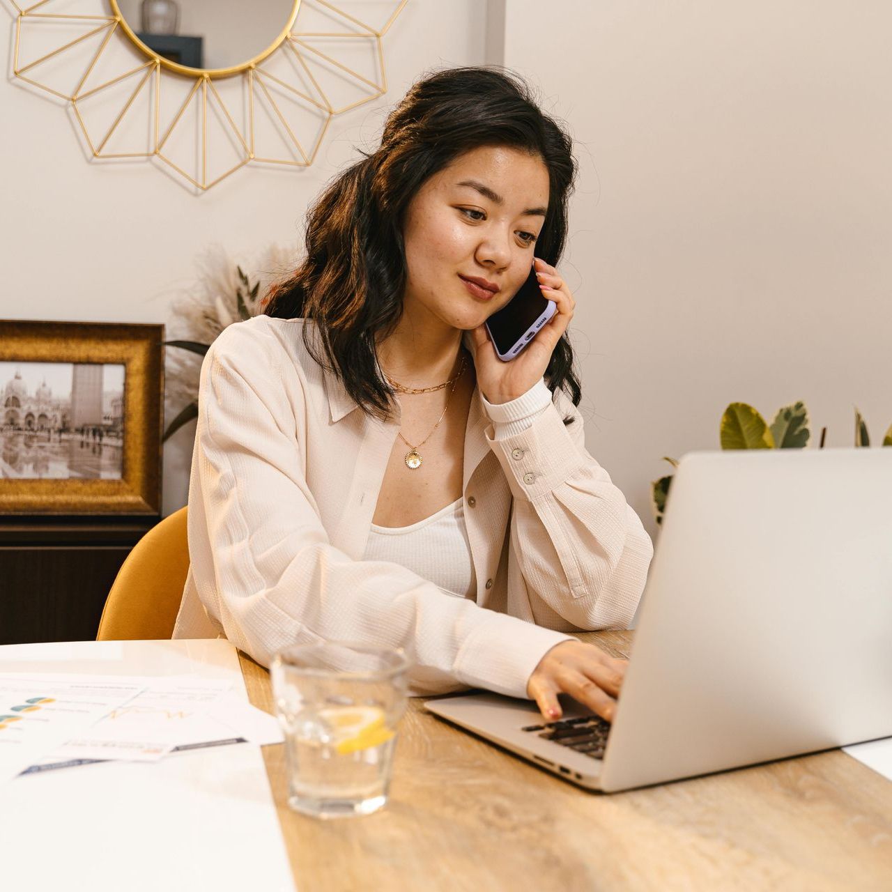 A woman is talking on a cell phone while using a laptop computer.