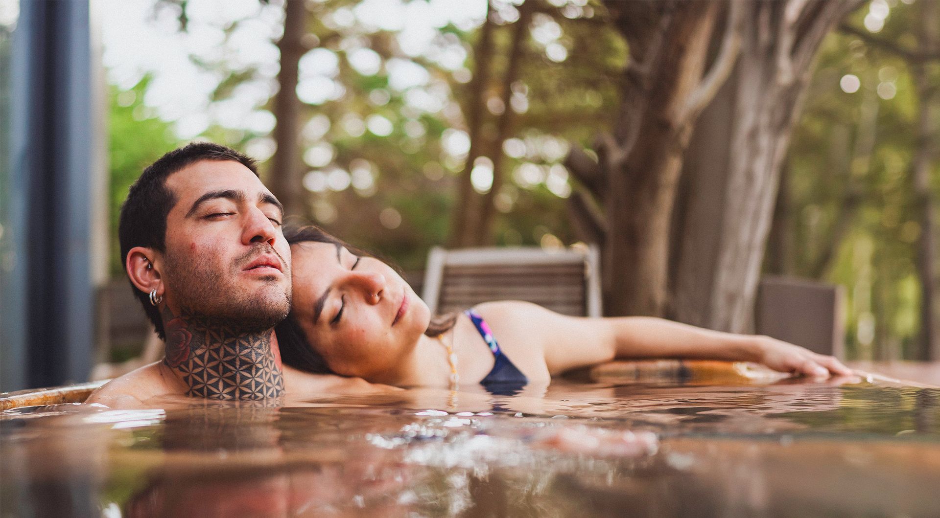 Dos personas relajándose con los ojos cerrados en un jacuzzi de madera al aire libre en medio de un fondo de bosque borroso.