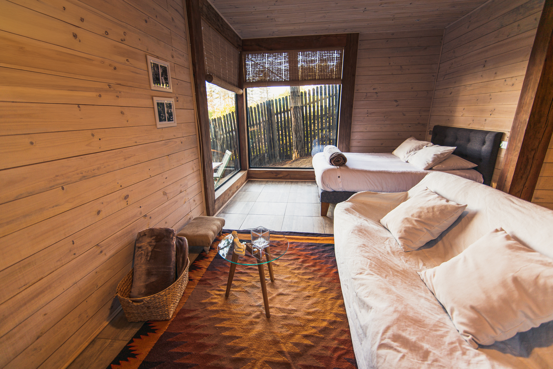 Acogedora habitación tipo cabaña de madera con dos camas, cesta de mimbre, alfombra y puertas de cristal que dan a un balcón.