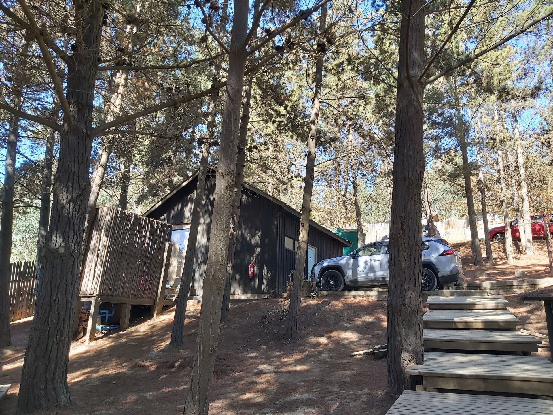 Cabaña de madera con coches aparcados y escalones de piedra en una ladera soleada.