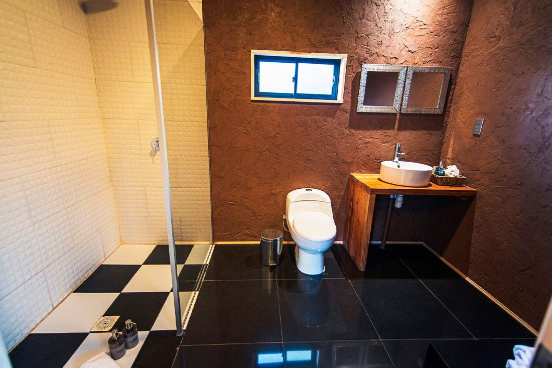 Baño moderno con suelo de baldosas negras, inodoro blanco, lavabo con mueble y zona de ducha alicatada.