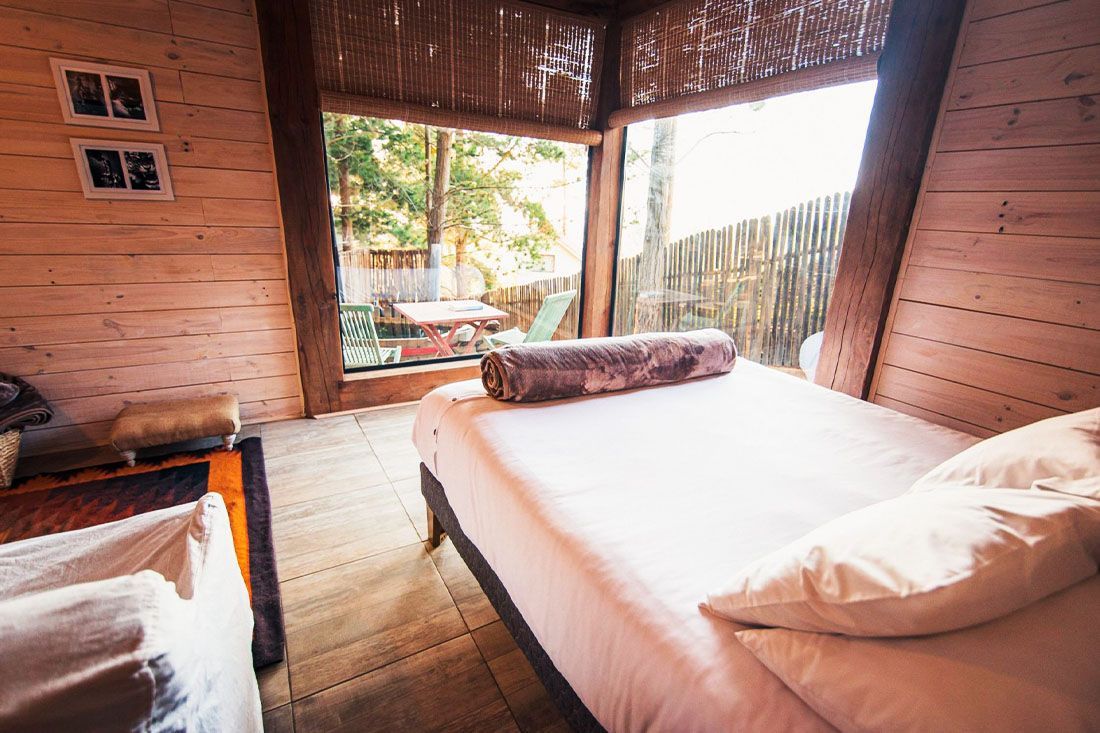 Acogedora habitación tipo cabaña de madera con dos camas y una luminosa puerta abierta que da a un balcón tropical.