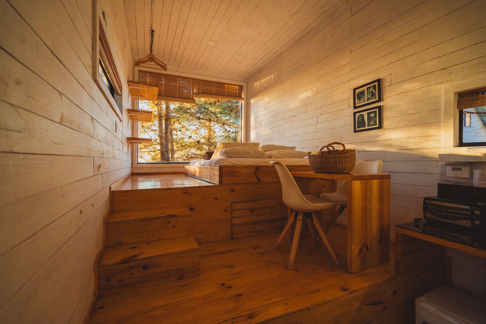 Interior acogedor de una cabaña de madera con cama alta, escritorio, silla y rincón soleado junto a la ventana.