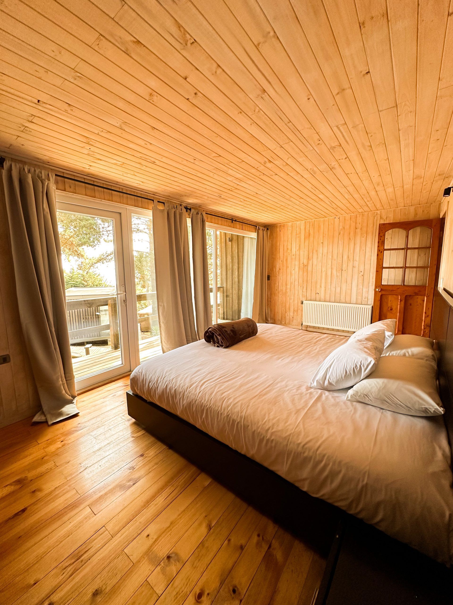 Acogedora habitación con paneles de madera, una cama grande, cortinas y puertas correderas que dan a un balcón.