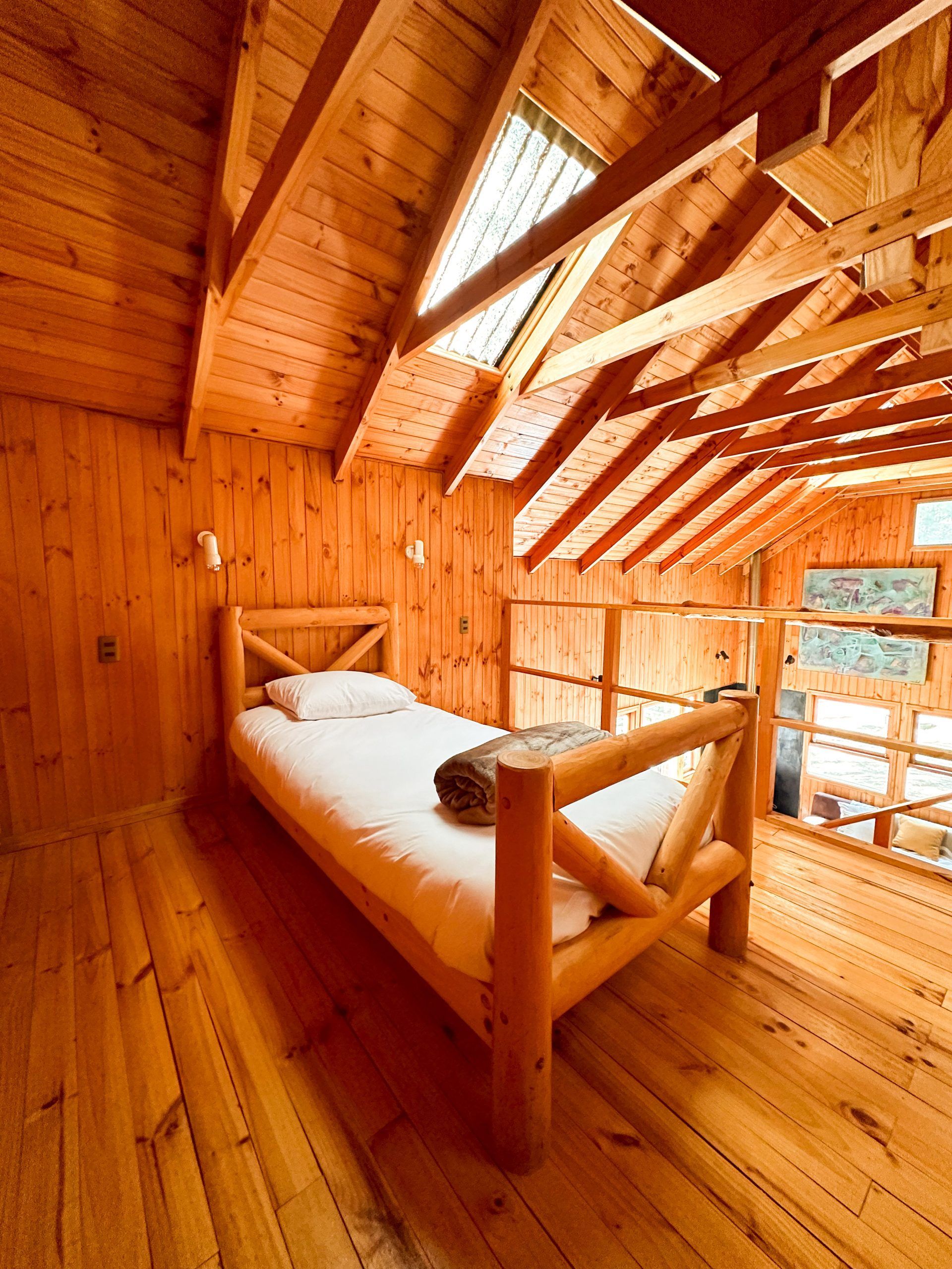 Habitación tipo loft de madera con cama individual, techo inclinado y ventanas de techo.