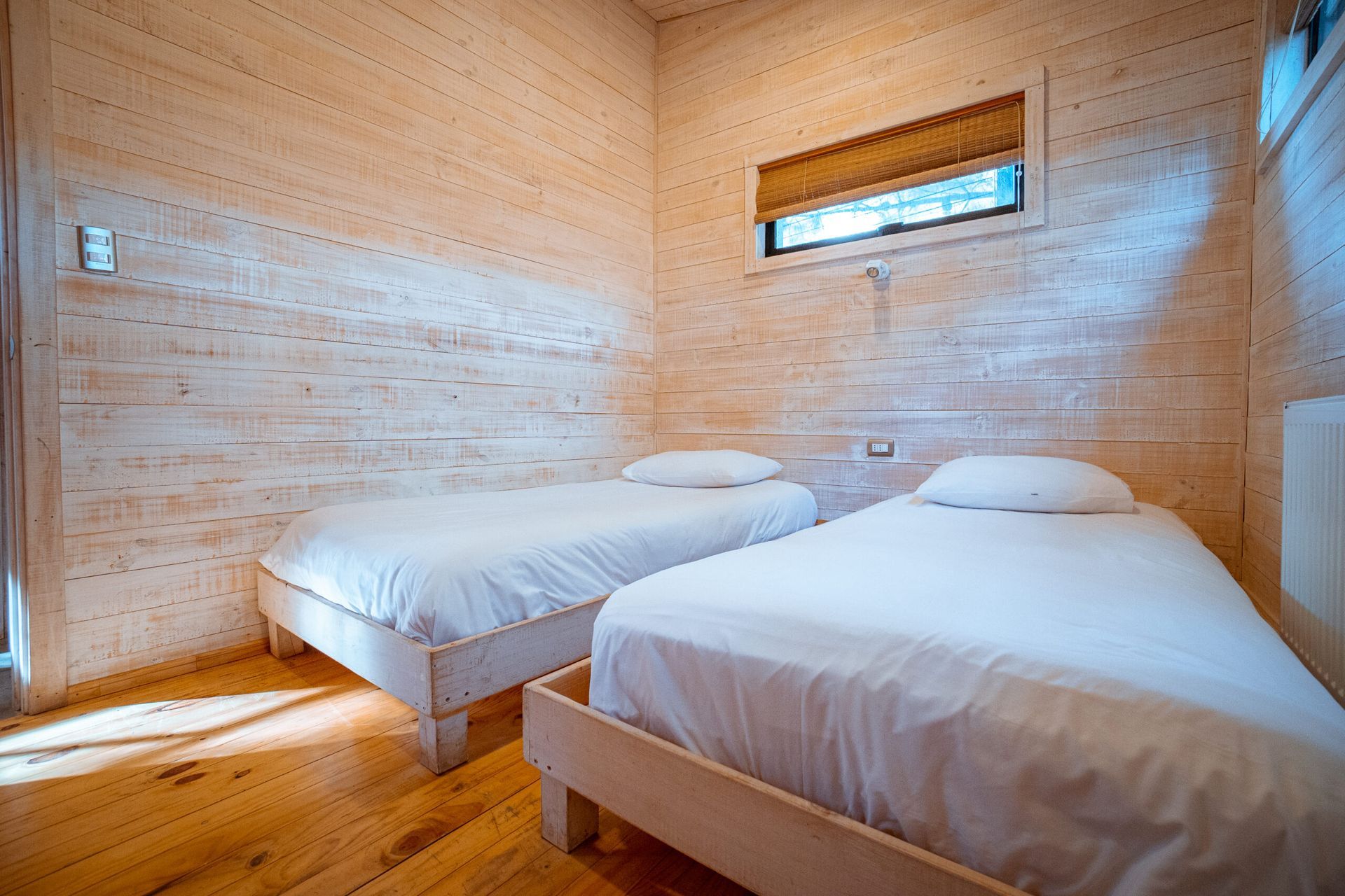 Habitación pequeña de madera con dos camas individuales, paredes de madera clara y una ventana estrecha.