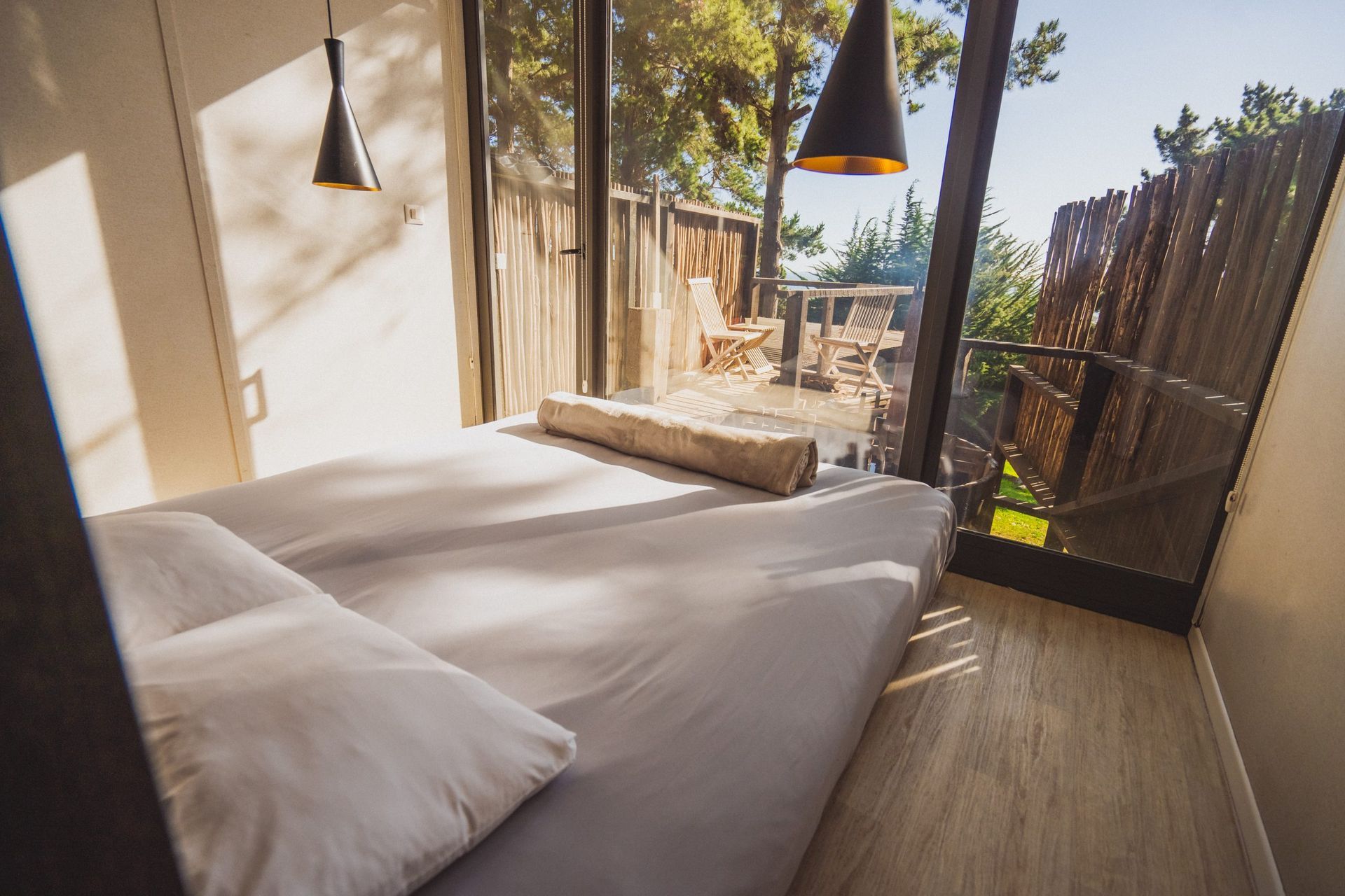 Acogedora cama de exterior tipo cabaña con cojines grises, almohada blanca y lámparas colgantes negras en un patio soleado.