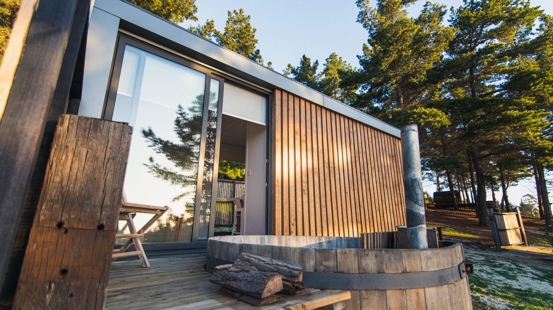 Una cabaña con revestimiento de madera y una puerta de vidrio, cuenta con un jacuzzi de madera en una terraza en un bosque.