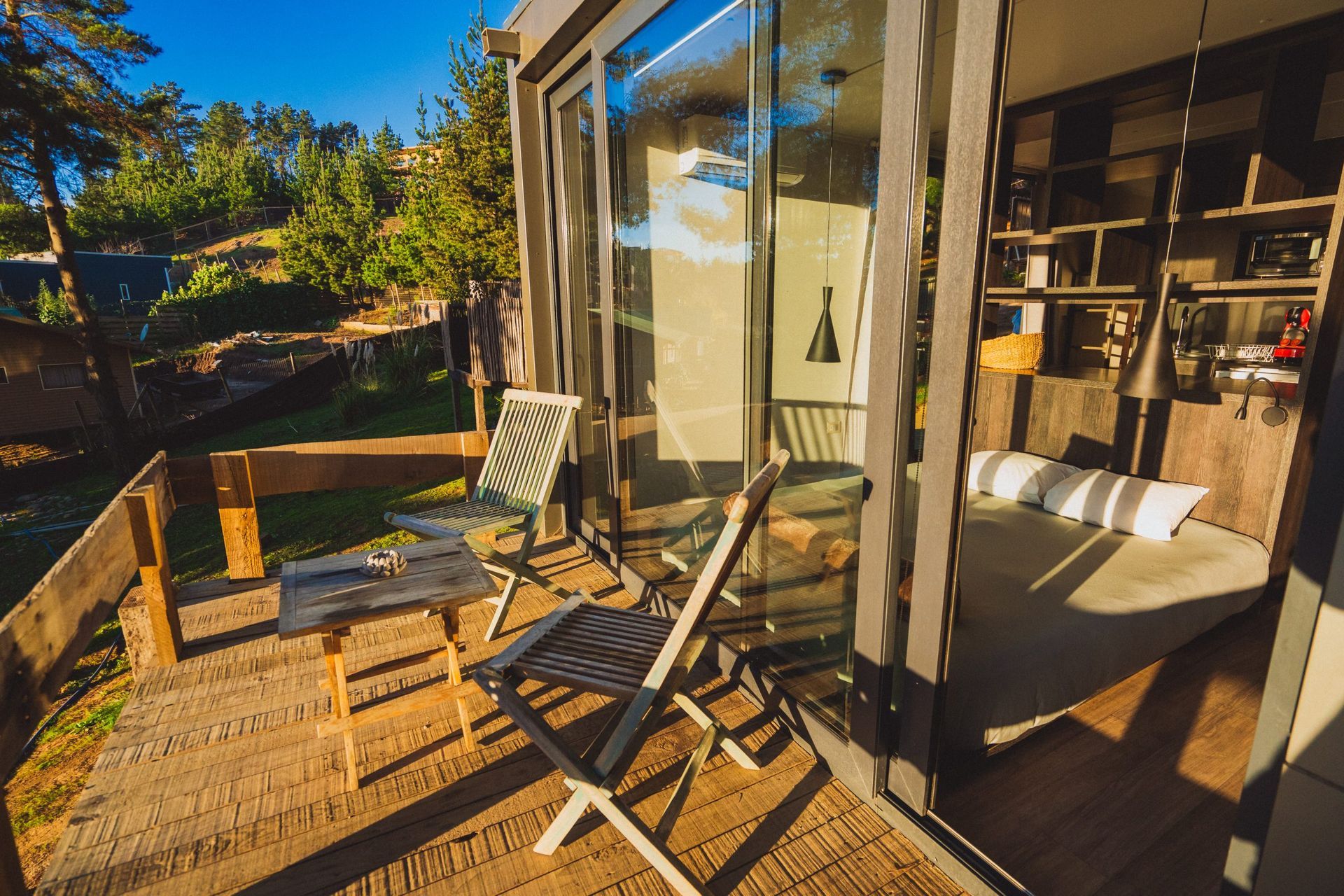 Un pequeño patio con una mesa de madera y dos sillas afuera de una cabaña moderna con vista a los pinos y un paisaje soleado.