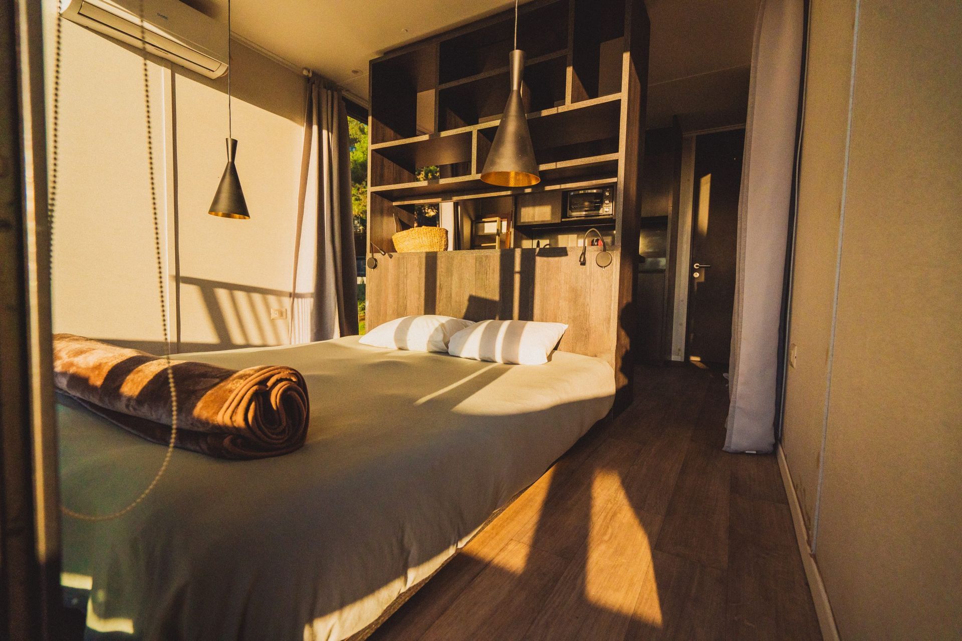 Un interior de dormitorio moderno con una cama, cálida luz solar, una estantería de madera y luces colgantes.