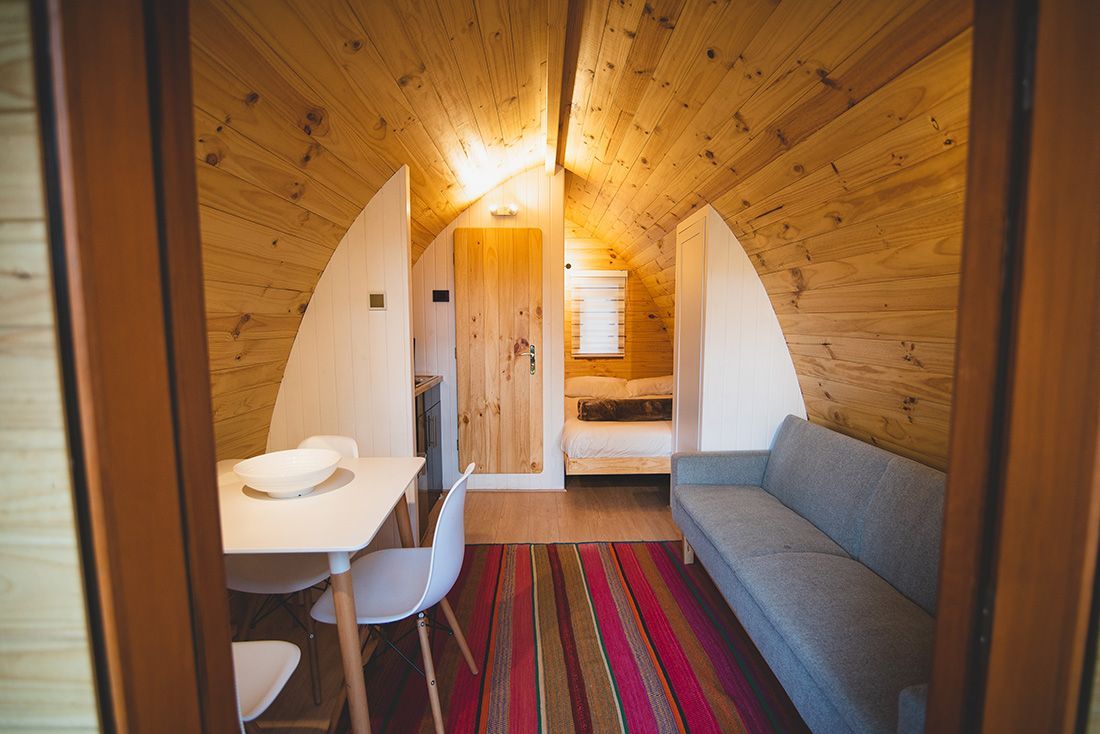 Interior de una pequeña cabaña con paneles de madera, que incluye una cama, un escritorio, un sofá y una alfombra a rayas.