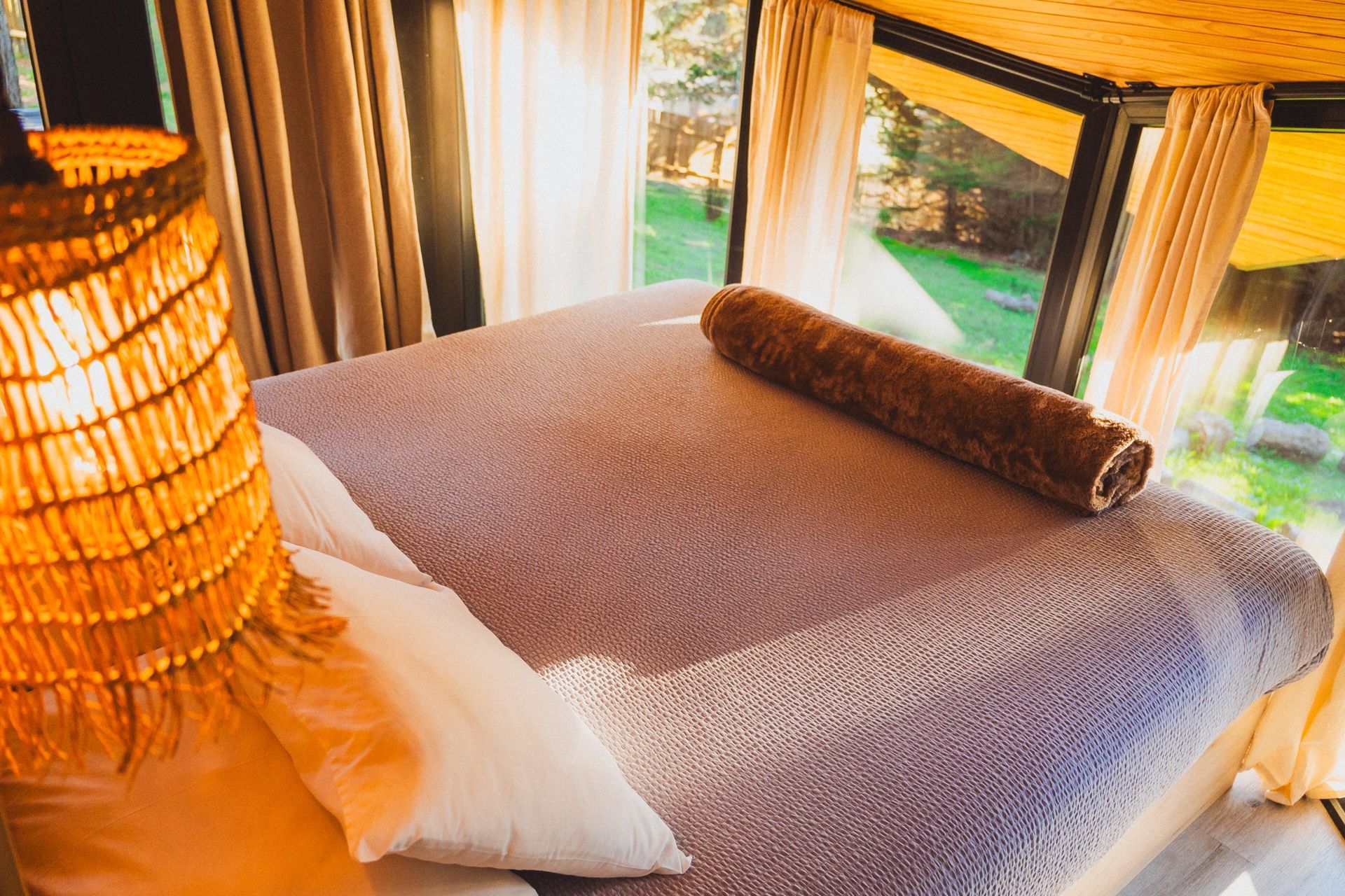 Cama acogedora con ropa de cama blanca y marrón junto a grandes ventanales con vistas a la vegetación y la luz del sol.