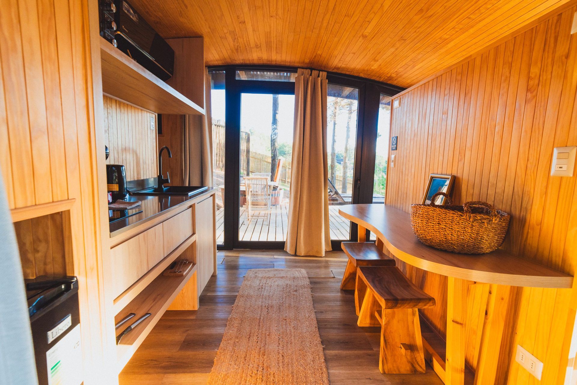 Cocina americana con paneles de madera, mesa estrecha, banco, cesta de mimbre y puertas de cristal que dan a una terraza.