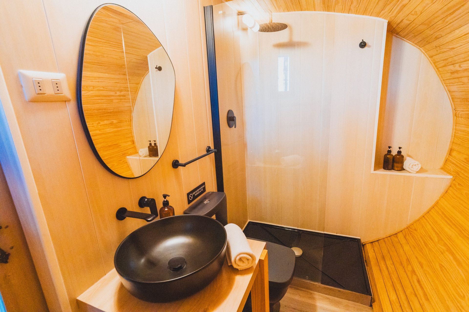 Baño pequeño con paredes revestidas de madera, espejo redondo, lavabo negro y puerta de ducha curva.