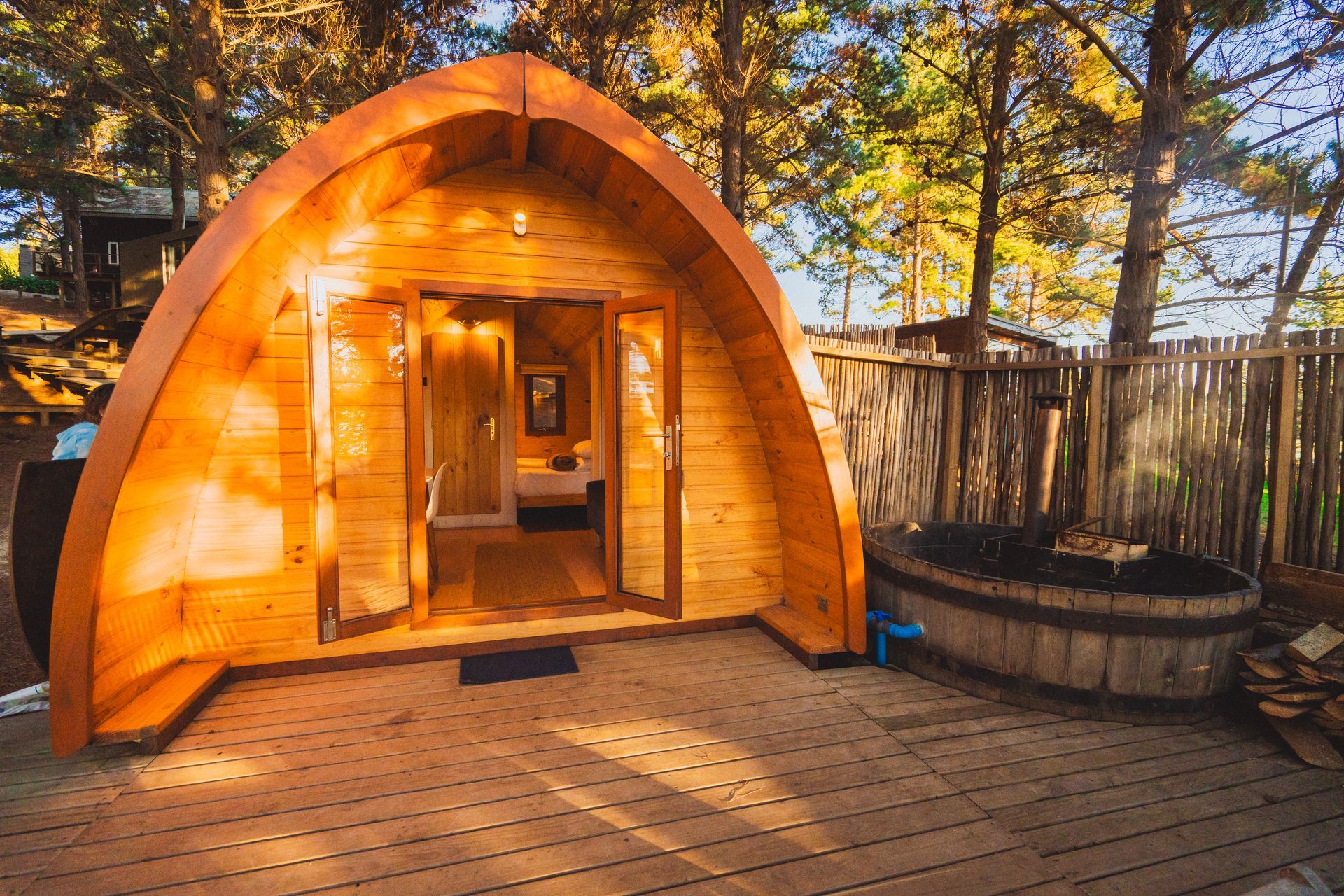 Sauna de barril de madera con puerta abierta en una terraza en un entorno boscoso al aire libre.