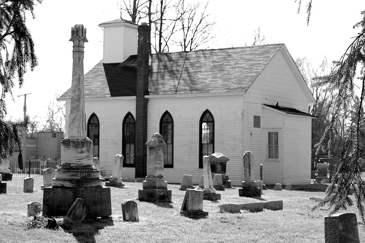 Black-and-white photo of a small cemetery beside a stone church with arched windows and gravestones