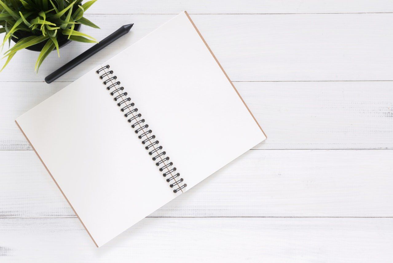 Open spiral notebook on white wooden desk with a green plant and black pencil