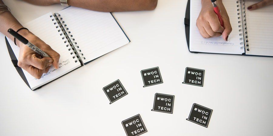 Hands writing in notebooks around scattered black tokens on a white table