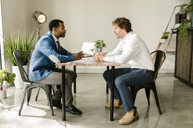Two people meeting at a small table in a bright office, talking over coffee and papers.