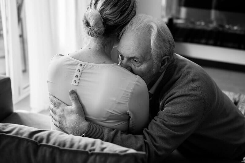 Two people embrace on a couch in a softly lit room, one comforting the other.
