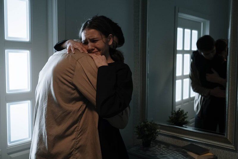 Two people hugging by a window, reflected in a wall mirror in a dim room.