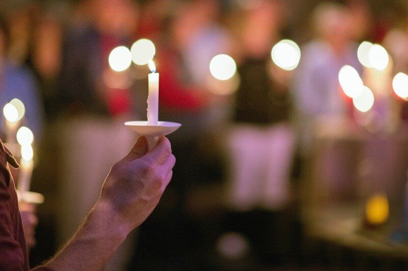 Hand holding a lit candle during a candlelight vigil, with blurred people and lights in the background