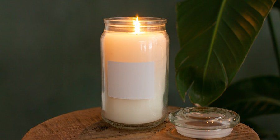 Lit white candle in glass jar beside a small glass lid on a wooden table with a green leaf background