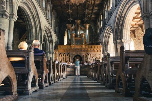 Inside a grand cathedral, a person stands in the aisle beneath tall arches and ornate chandeliers.