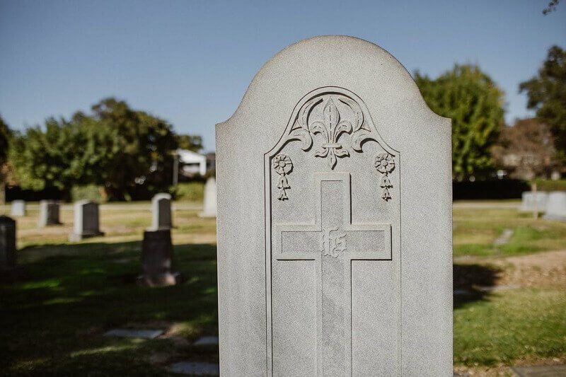 White gravestone with cross engraving in a cemetery, with trees and other headstones in the background